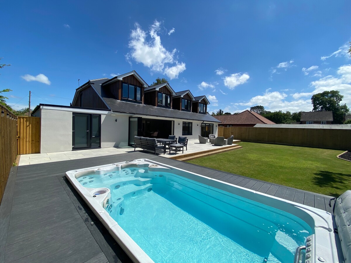 The exterior of the property is showcased with a spacious garden and a hydropool swim spa. The large house features multiple pitched roofs, with outdoor seating arranged on a patio area, surrounded by well-maintained grass and a clear blue sky.