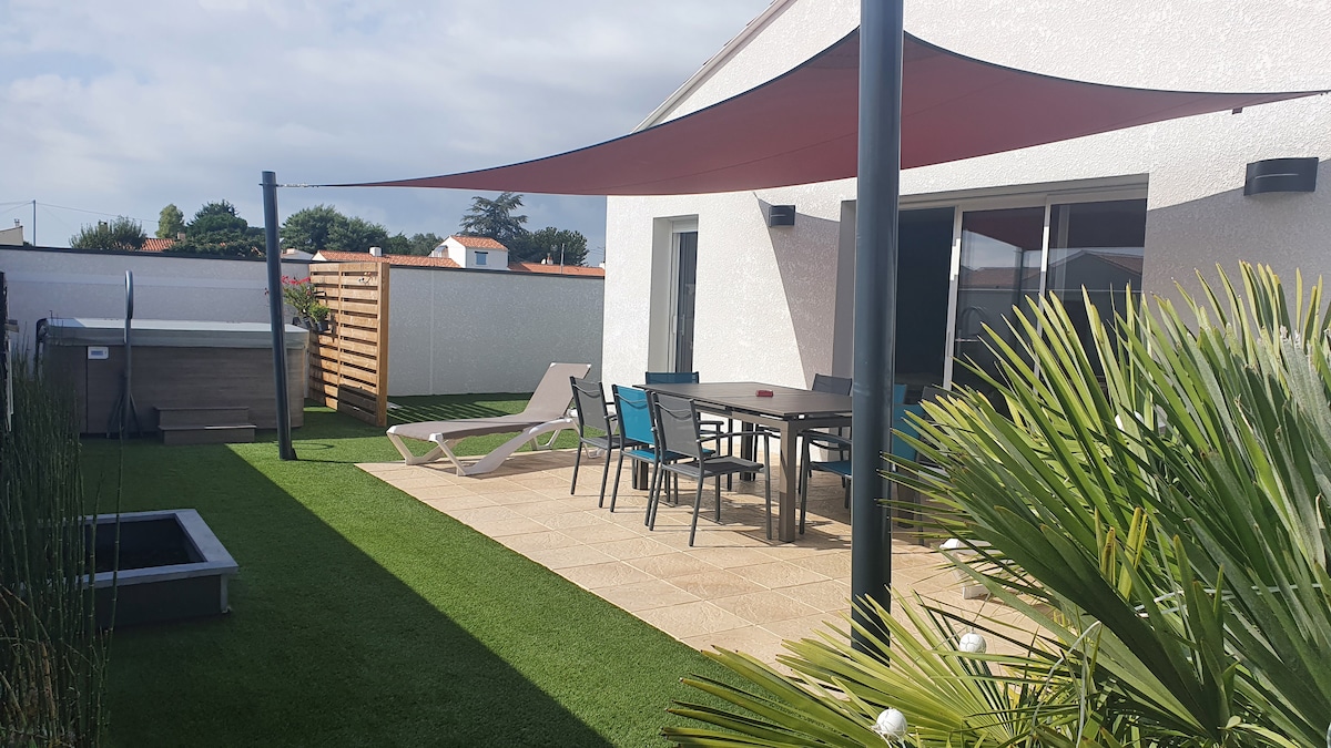 An outdoor terrace is displayed, featuring a shaded area with a table and chairs. Two lounge chairs are positioned alongside a neatly landscaped garden, which includes a small, rectangular outdoor pool. A privacy screen and storage unit are visible in the background.