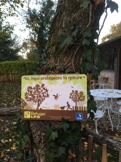 A sign affixed to a tree informs visitors about the conservation efforts of the refuge LPO. The illustration features trees and wildlife, complementing the natural surroundings. The area is framed by a wooden fence and a small outdoor seating arrangement, enhancing the peaceful ambiance.