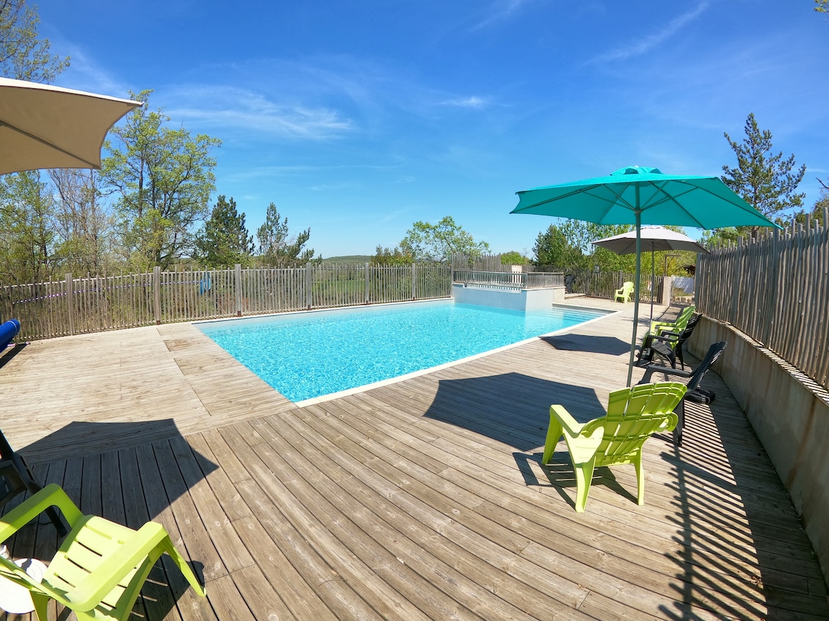 A serene swimming pool is surrounded by a wooden deck, featuring vibrant green lounge chairs. Large umbrellas provide shade, while trees and a gentle landscape create a calm backdrop. The water's clear surface reflects the bright blue sky, inviting relaxation.