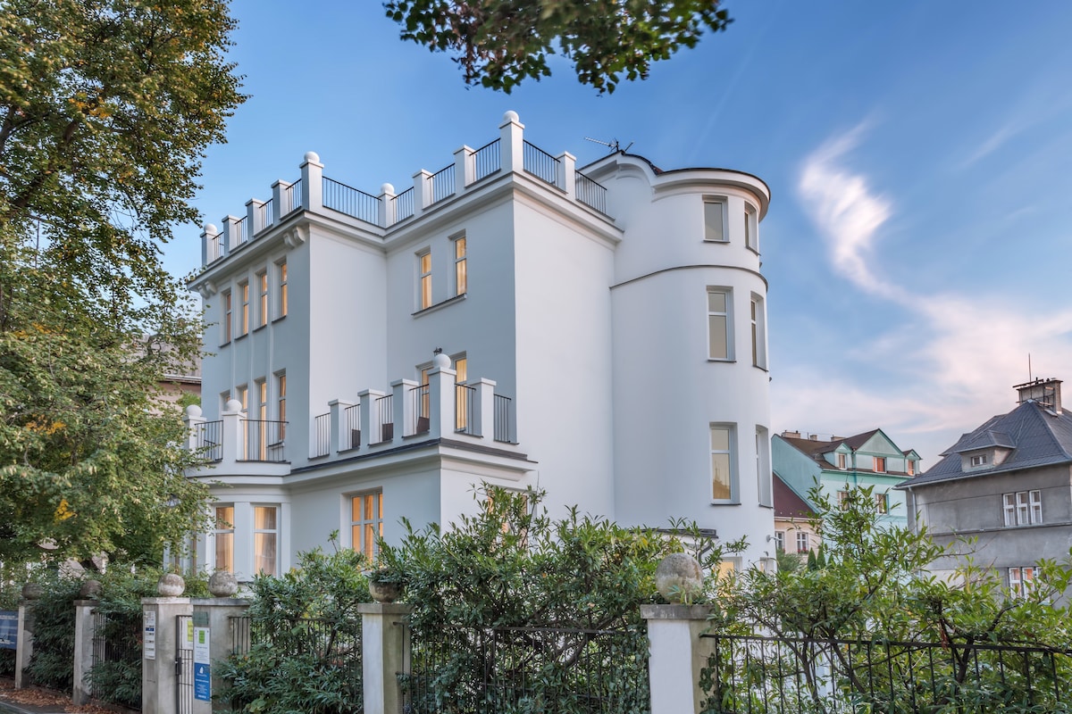 The exterior of a modern, multi-story villa is showcased, featuring a smooth white facade and multiple balconies. Surrounding greenery adds a natural touch, while large windows allow for ample natural light. The structure is framed by clear blue skies, enhancing the welcoming appearance.