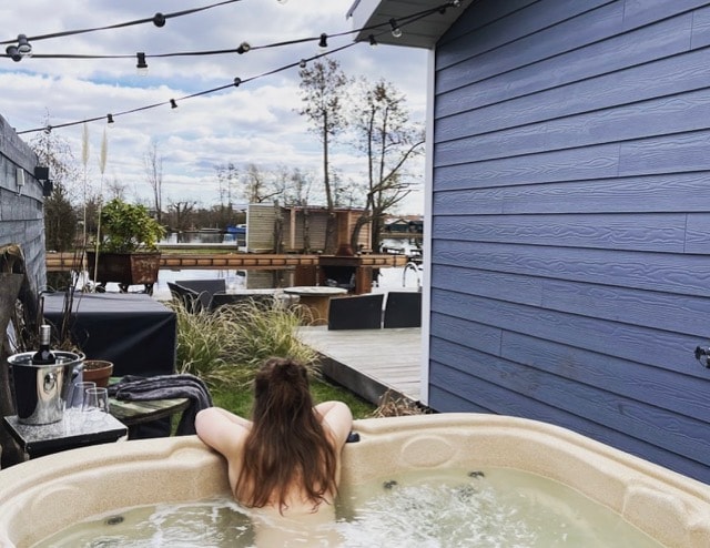A private jacuzzi is visible at the outdoor space, surrounded by landscaped areas and wooden decking. Soft lighting from string lights creates a warm ambiance. The calm water reflects the sky, while outdoor seating and plant life can be seen in the background, enhancing the relaxation setting.