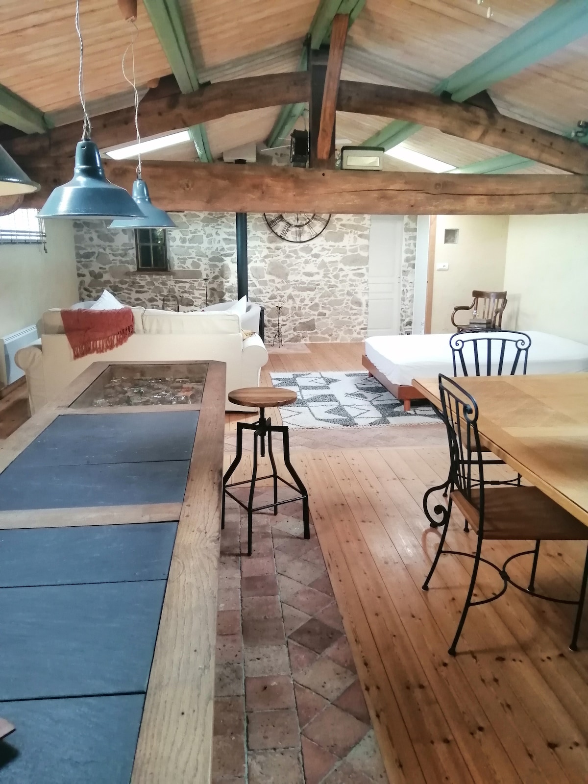An open living space features wooden beams and a high ceiling, complemented by exposed stone walls. A comfortable seating area includes a light-colored sofa and a patterned rug. A dining area with a wooden table and chairs is visible, enhancing the cozy atmosphere.