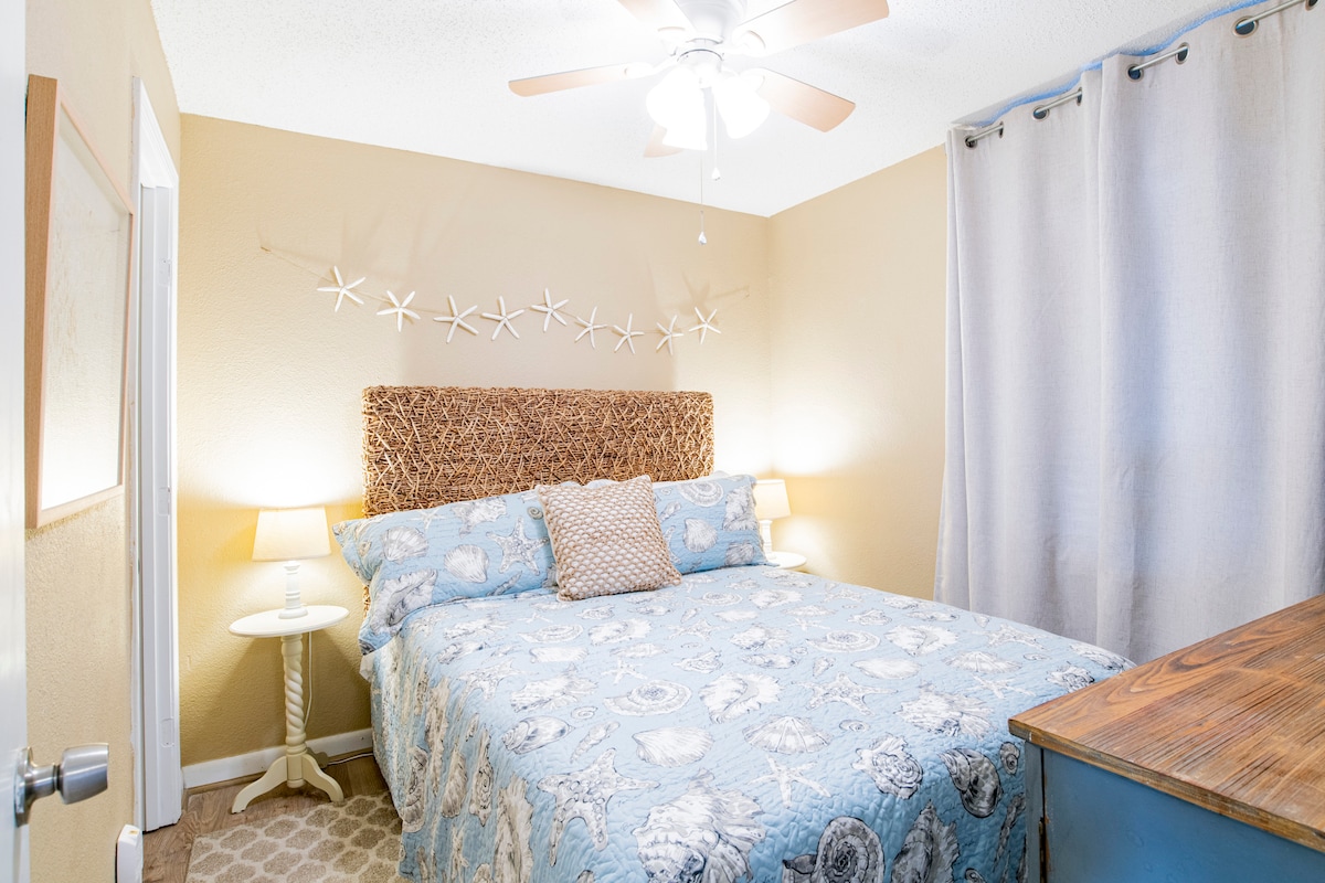 นี่คือห้องนอนด้านหลังที่แสดงการตกแต่งชายหาดที่น่ารักที่นอนสบายและคุณสามารถเห็นพรมข้างเตียงโต๊ะข้างเตียงและโคมไฟสำหรับแต่ละคน 