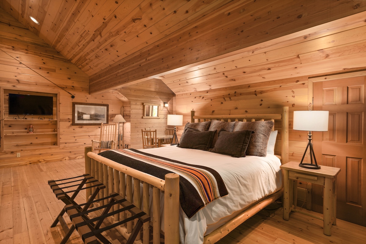 A spacious room features a wooden ceiling and walls, contributing to a rustic atmosphere. A large bed with a patterned throw is complemented by bedside tables and lamps. Two rocking chairs provide additional seating, while a flat-screen TV is mounted on the wall.