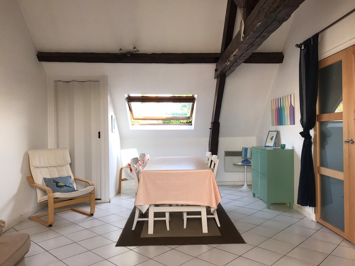 A light-filled dining area features a large table surrounded by white chairs. A wooden beam crosses the ceiling, complementing the minimalist decor. A green cabinet and a small desk are positioned against the wall, with a window above providing natural light.