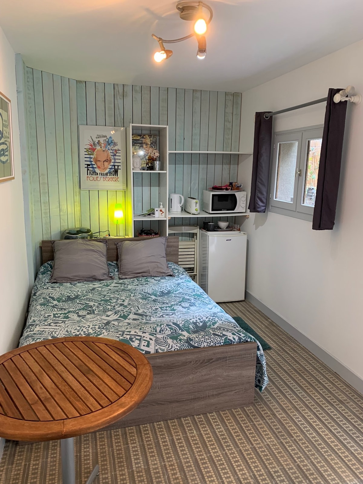 A cozy sleeping area features a spacious bed with a patterned coverlet and two decorative pillows. A small table is positioned nearby. A kitchenette with a refrigerator and microwave is visible on the wall, complemented by soft lighting and a piece of wall art.