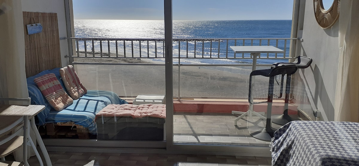 A spacious terrace with a view of the sea features a comfortable seating area, including lounge chairs and a small table. Natural light floods the space, creating a bright atmosphere, while the ocean can be seen reflecting the sunlight in the distance.
