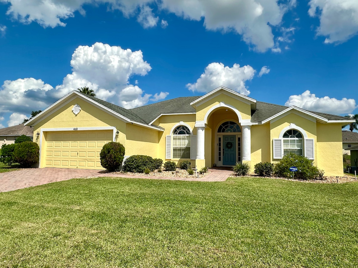 The exterior of a bright yellow villa is featured, accented by a well-maintained lawn and landscaped garden. A spacious driveway leads to a two-car garage, with arched windows enhancing the front façade. Fluffy clouds are visible in the blue sky above.