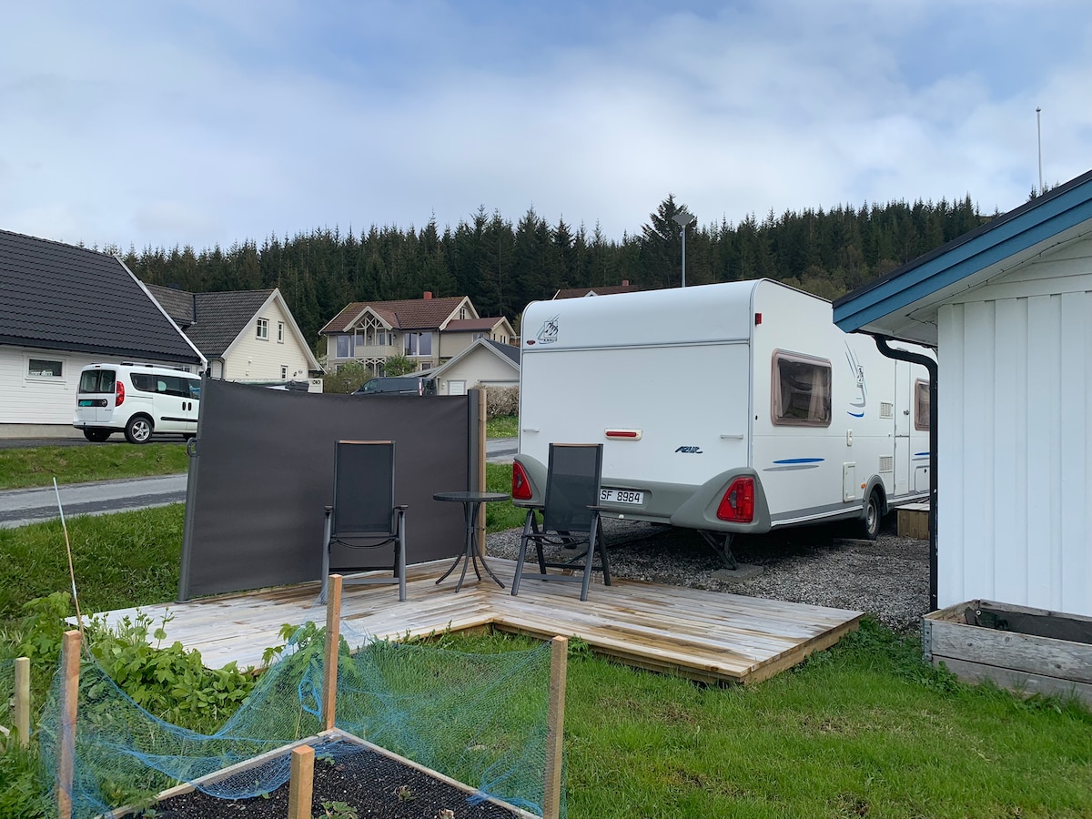 The image displays a camping caravan parked next to a wooden deck with two outdoor chairs and a small table. A privacy screen is positioned nearby. Rolling green hills and scattered houses are visible in the background, set against a cloudy sky.