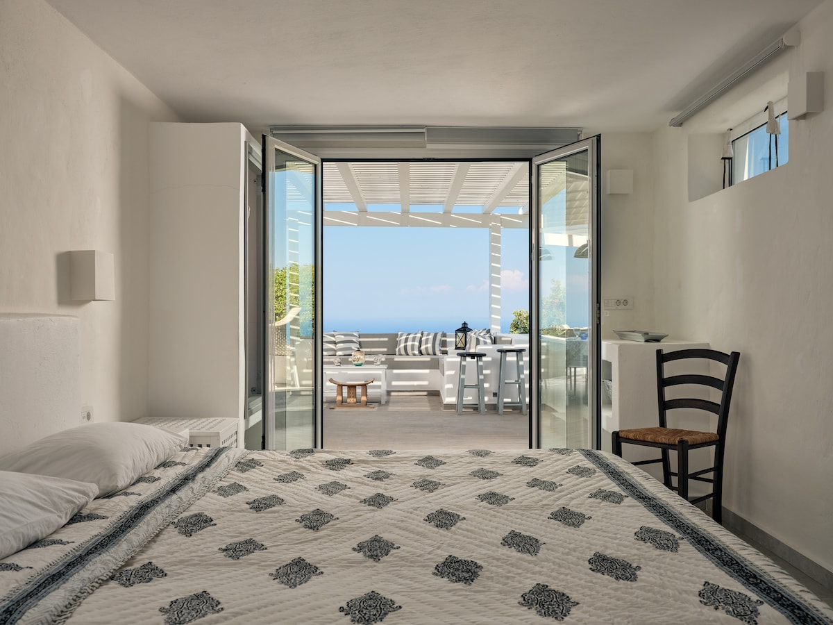 A spacious sleeping area features a white quilted bedspread with subtle patterns. Large glass doors open to a private terrace with outdoor seating, offering views of the surrounding landscape and sea. A chair is positioned near the bed, enhancing the room's functionality.