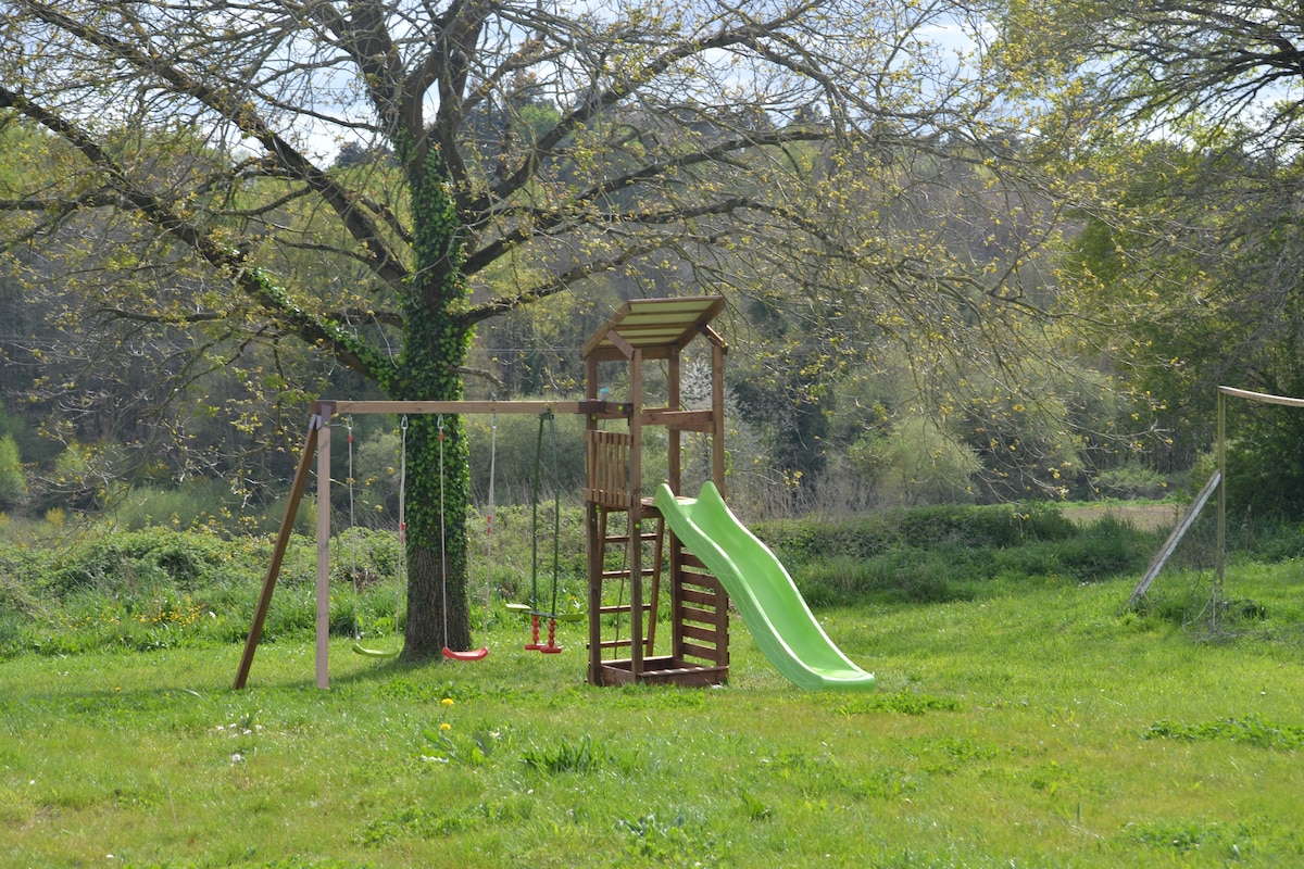 A children’s play area features a wooden playset with a yellow slide and swings. The playset is surrounded by greenery, including a large tree and an open grassy space, creating a serene outdoor environment.