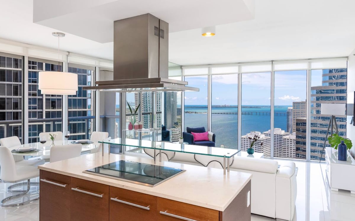 An airy kitchen and living area are shown, featuring a large island with a stovetop. Floor-to-ceiling windows offer expansive views of the bay, while modern, white dining chairs surround a round table. The sleek design emphasizes natural light and the scenic backdrop.