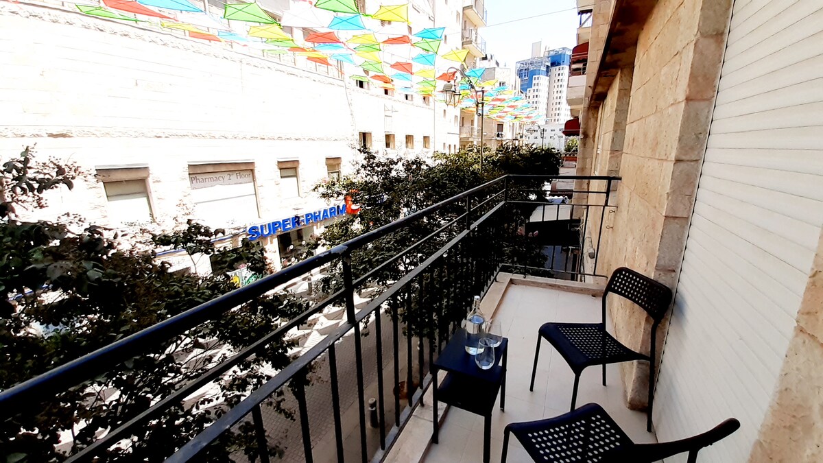 A small balcony is visible, furnished with two black chairs and a glass table. Colorful fabric canopies are draped above the street below, creating an engaging overhead display. The view includes nearby urban buildings and greenery from potted plants along the balcony railing.