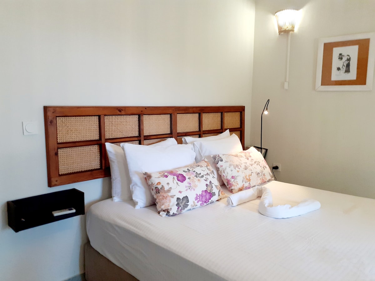 A calming sleeping area is showcased, featuring a spacious bed adorned with white linens and decorative pillows with floral patterns. A bedside table is positioned next to the bed, complemented by a wall lamp that provides soft lighting. Natural tones enhance the overall serenity of the space.