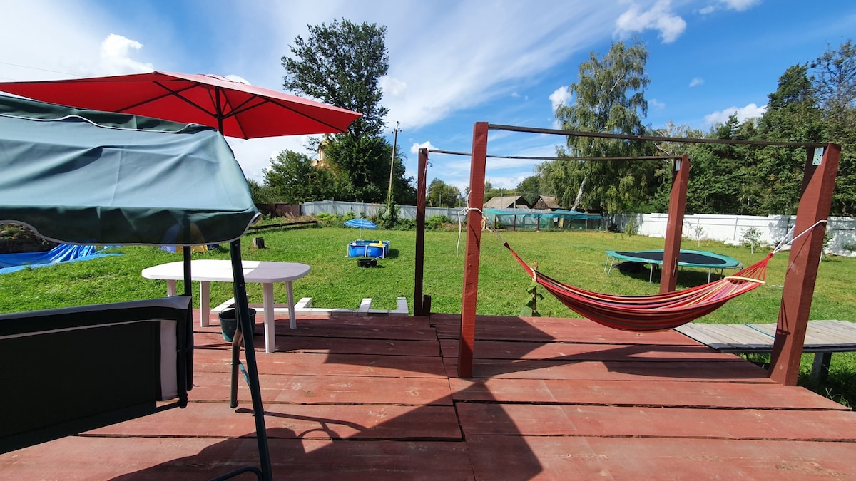 A spacious outdoor deck is shown, featuring a hammock and a table with chairs shaded by an umbrella. The green lawn is visible in the background, along with a trampoline and various residential structures partially obscured by trees. A clear blue sky is present above.