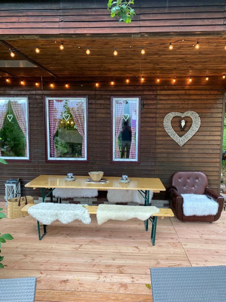 An outdoor seating area features a long wooden table accompanied by benches adorned with soft coverings. A brown armchair sits nearby, providing additional comfort. String lights illuminate the space, creating a cozy ambiance, while patterned curtains frame the windows in the background.