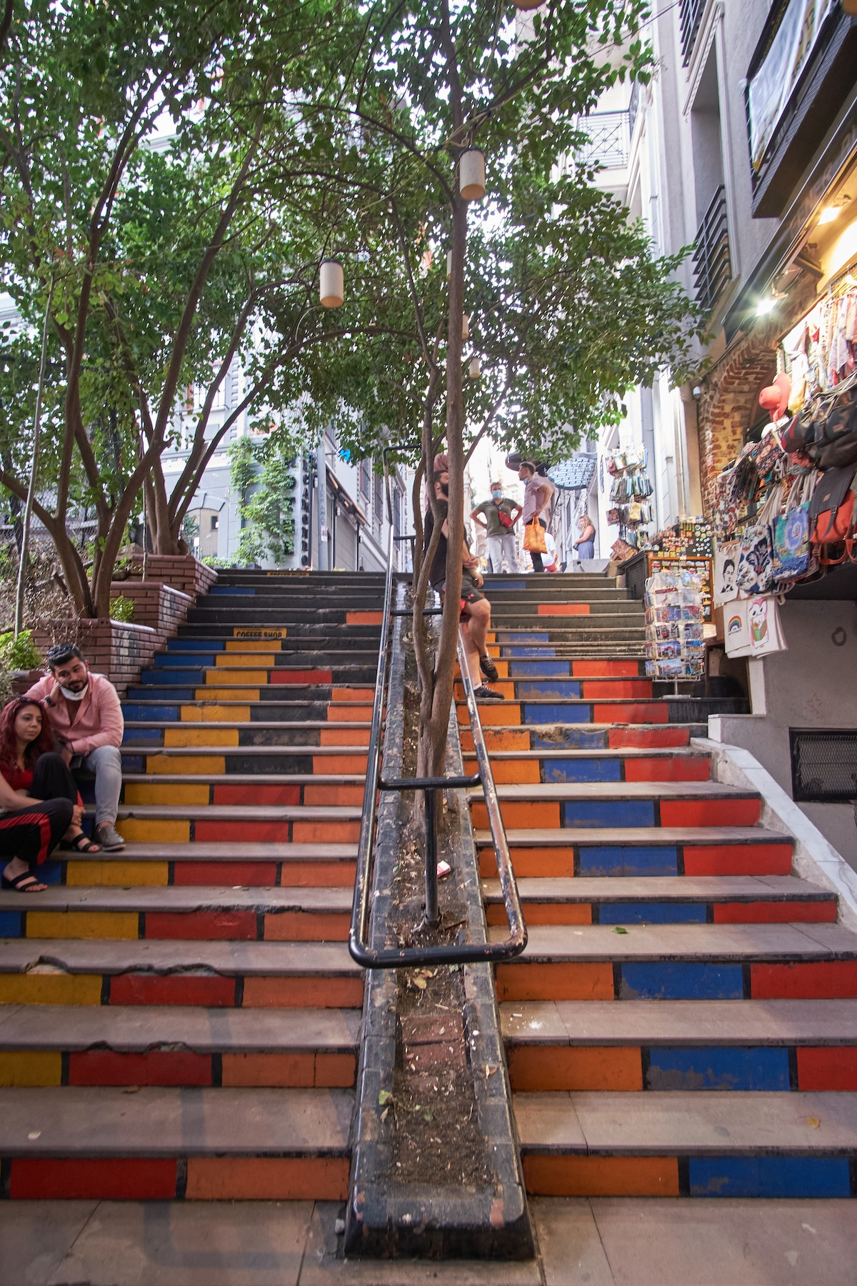 A set of stairs is decorated with colorful stripes in red, blue, and yellow. Green trees provide foliage along the pathway, and small white lights hang from branches, creating an inviting atmosphere. Vendors and visitors can be seen in the background, adding life to the scene.