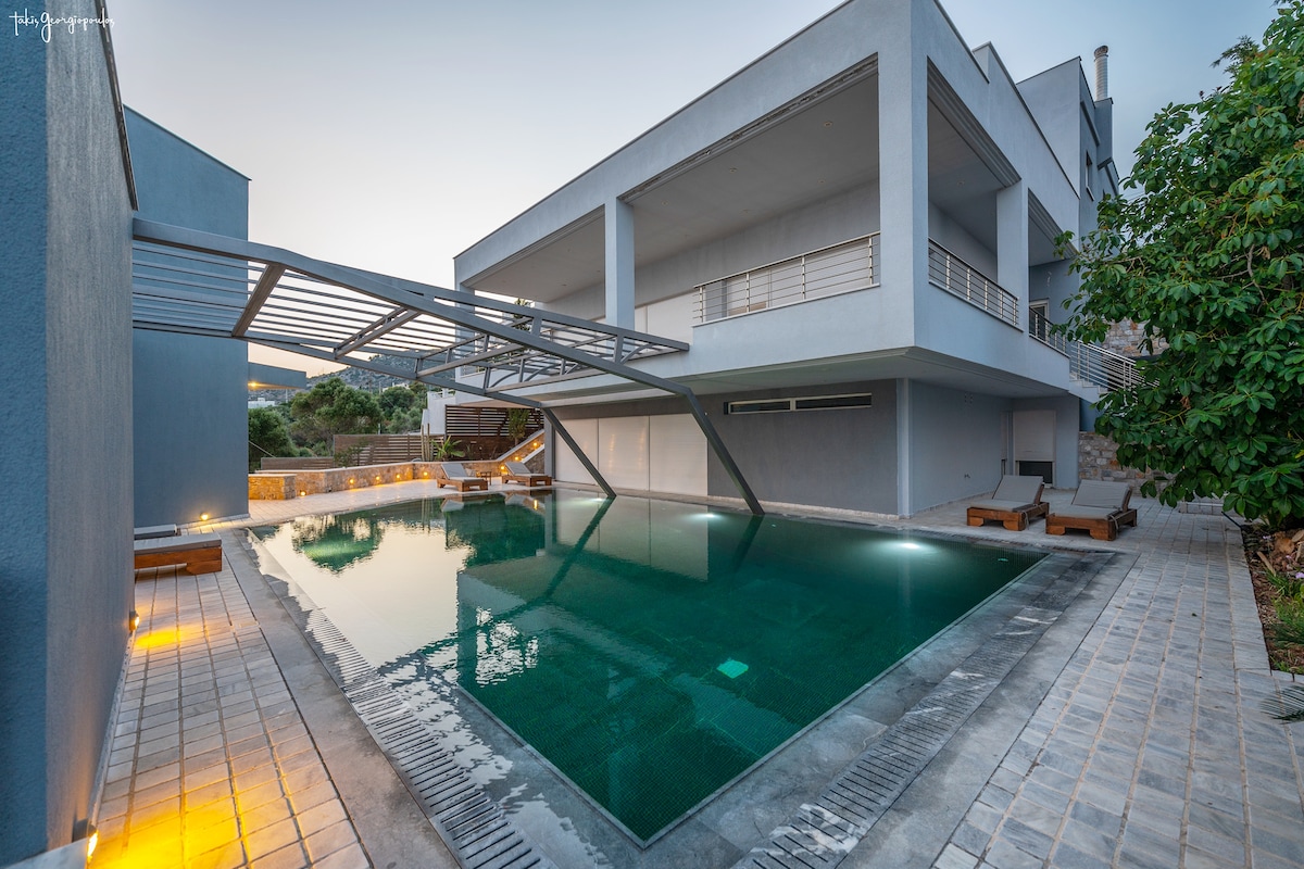 A modern swimming pool is situated within a contemporary architectural setting, featuring clear water and a sleek design. The pool area is surrounded by smooth tiles and a landscaped garden, with soft lighting illuminating the space as evening approaches.