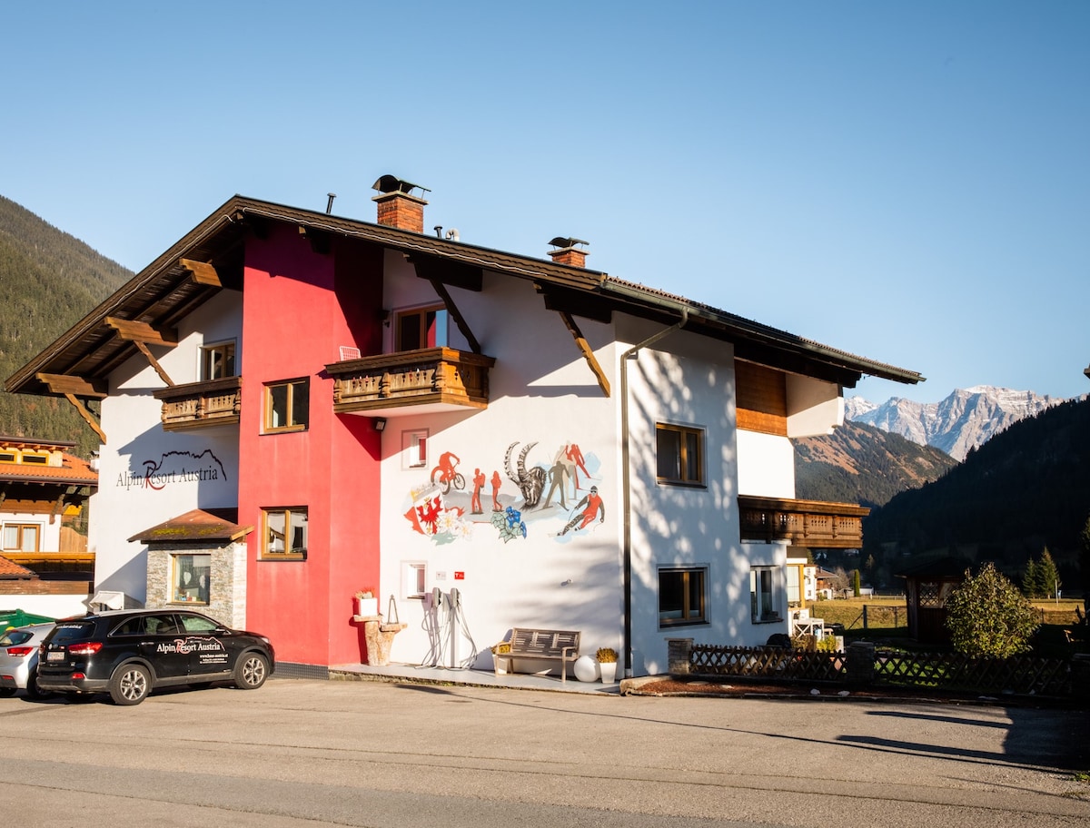 A charming two-story building is presented, featuring a mix of red and white exterior walls. Artistic murals decorate the façade, and surrounding mountains provide a scenic backdrop. A small seating area with benches and a parking area are visible in the foreground.