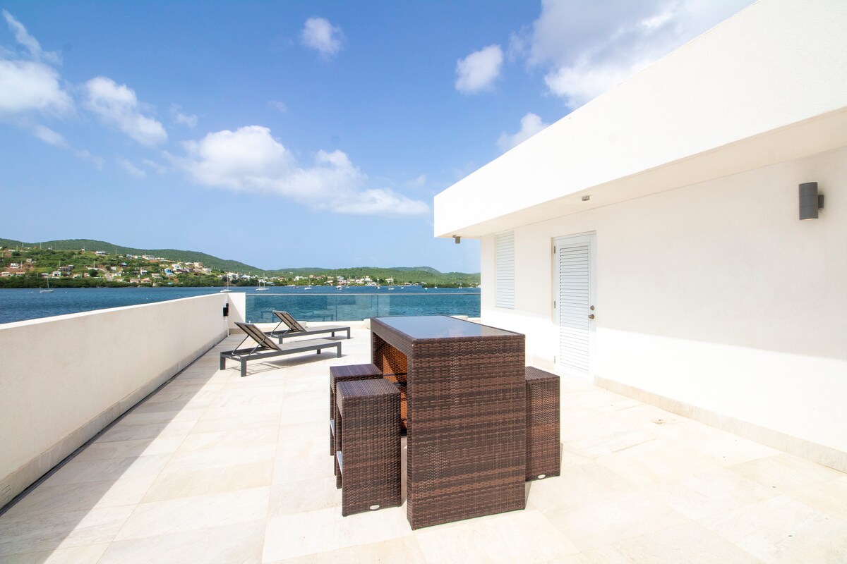 An outdoor rooftop space is presented, featuring a dark wicker dining set accompanied by matching chairs. Two lounge chairs are positioned nearby, with panoramic views of the water and surrounding landscapes. Clear skies and soft clouds complement the tranquil atmosphere.