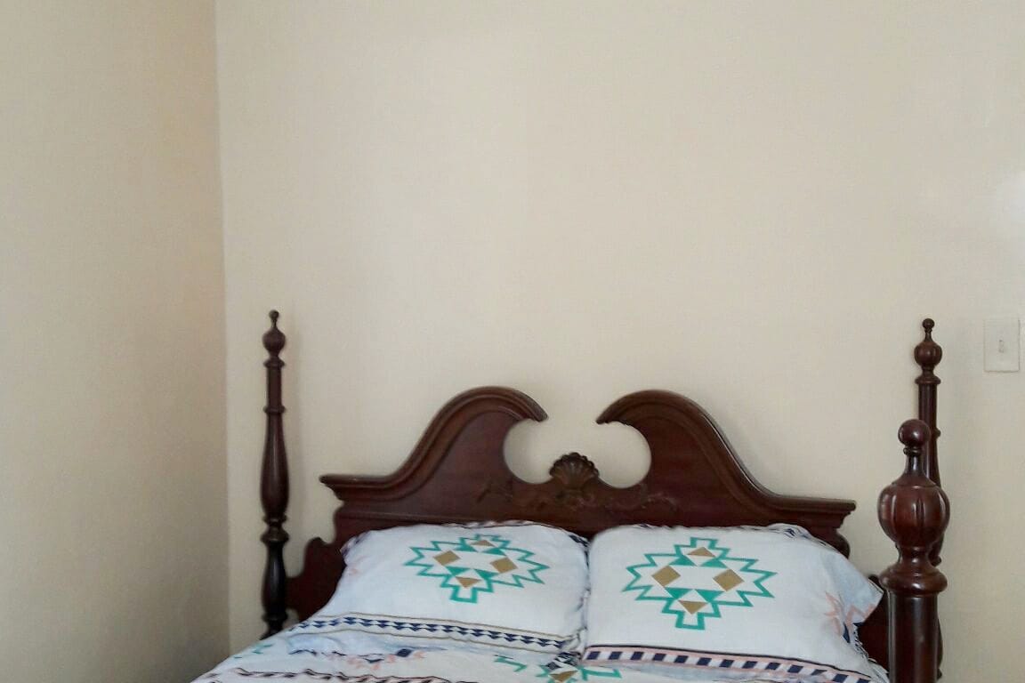 A wooden bed frame with decorative posts is positioned against a light-colored wall. The bed features a fitted sheet adorned with geometric patterns, complemented by two matching pillows. Natural light enhances the calming atmosphere of the room.