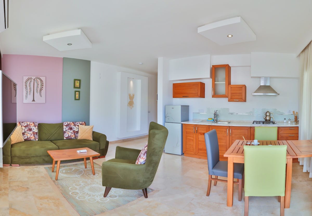 An open living area features a green sofa with decorative pillows, a light wood coffee table, and an adjacent dining table with chairs in various colors. The kitchen, equipped with wooden cabinets and modern appliances, is visible in the background. Large windows allow natural light to brighten the space.