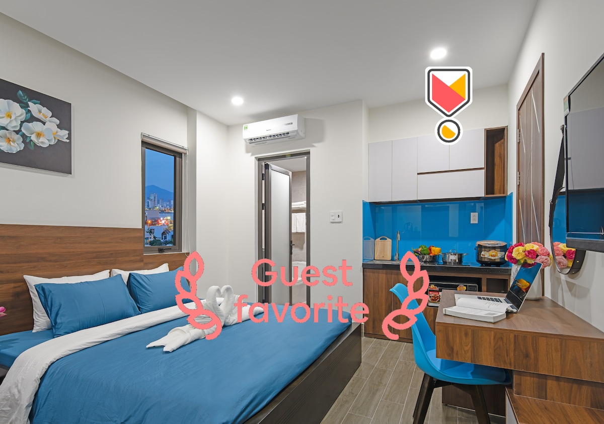A compact room features a spacious bed dressed in blue linens, accompanied by a small table and blue chair. Natural light enters through the window, offering a view of the city. A kitchenette is visible, equipped with essential appliances. Artwork with floral designs adorns the wall.