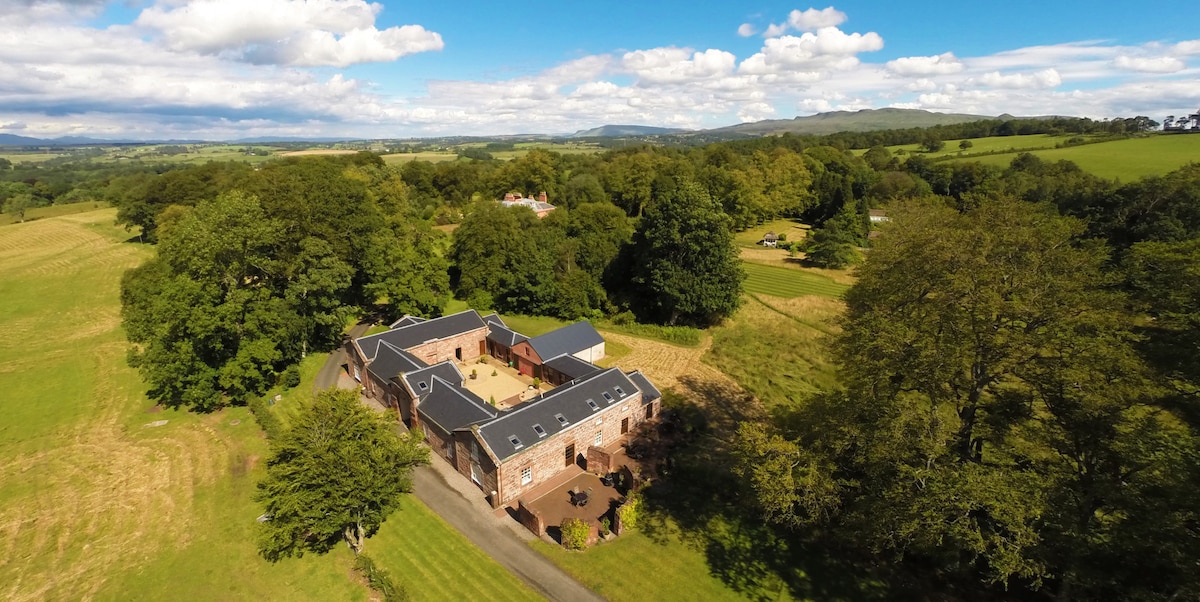An aerial view captures Sycamore Cottage, showcasing a rustic building surrounded by lush greenery and open fields. The charming exterior features a combination of brick and modern roofing, providing a peaceful atmosphere complemented by the natural landscape.