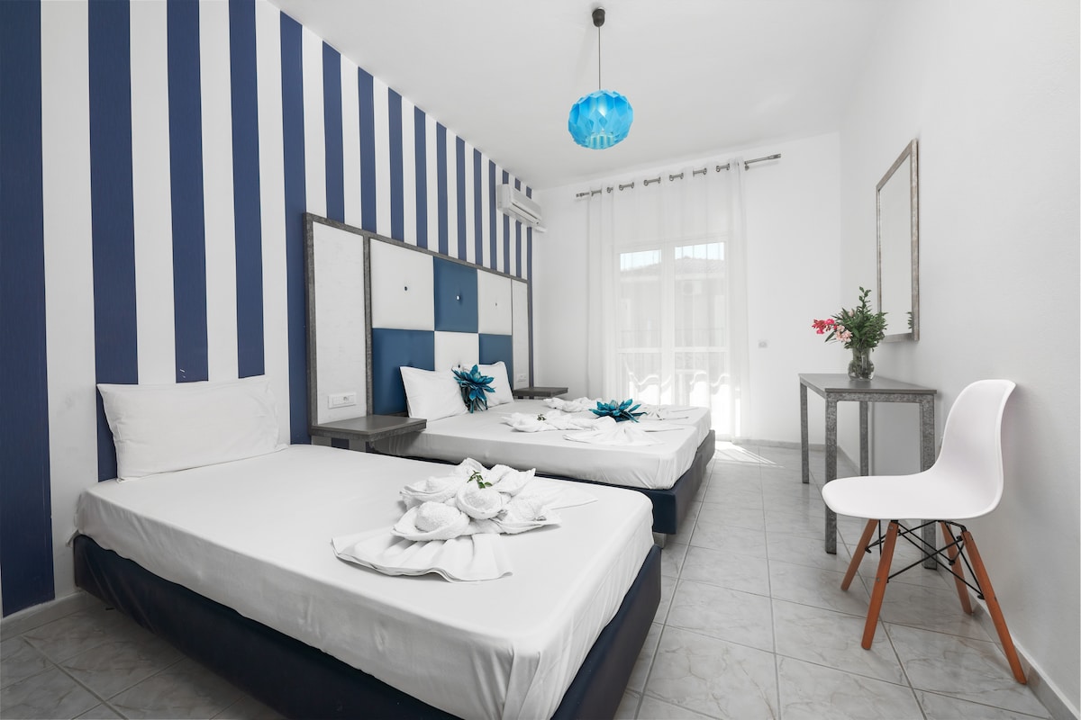 A well-maintained room features blue and white striped walls with two spacious beds made up with white linens. A small table with a mirror and a decorative vase sits nearby. Natural light enters through the large window, complemented by sheer curtains.