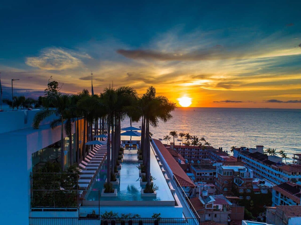 The rooftop features an infinity pool surrounded by palm trees, with lounge chairs arranged nearby. The sunset casts vibrant colors over the ocean, blending oranges and blues, while buildings in the background add to the coastal view of Puerto Vallarta.