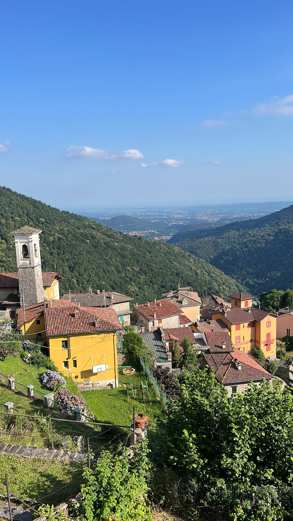 A panoramic view of a mountain village is presented, featuring traditional stone houses with terracotta roofs and colorful facades. Lush green hills rise in the background, and a church tower is visible, contributing to the scenic charm of the area. The landscape extends into the valley below.