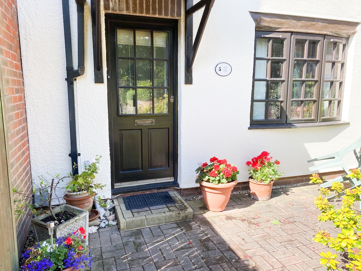 The entrance to the cottage features a dark wooden front door with a window above it. Flower pots containing vibrant red geraniums are positioned on either side, complementing the cottage's exterior. A paved path leads to the door, surrounded by well-maintained greenery.
