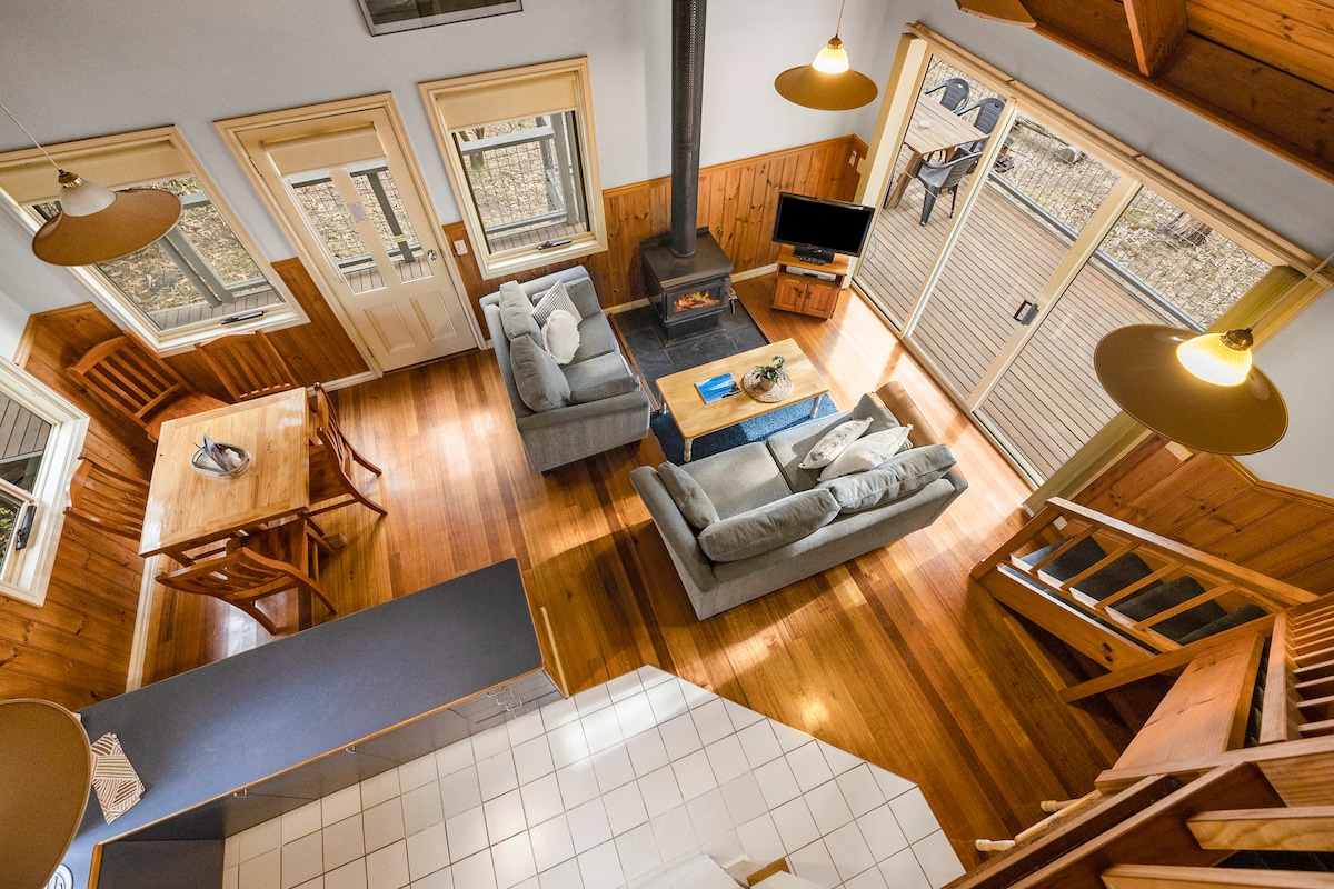 An overhead view of the living area showcases a spacious design with wooden accents. A comfortable seating arrangement surrounds a central wood-burning stove, while a dining table is positioned nearby. Large windows invite natural light, and a staircase leads to an upper level.