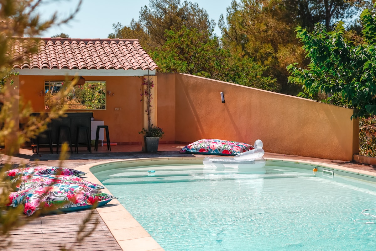 A refreshing swimming pool is framed by palm trees and a muted orange wall. Colorful inflatable loungers rest on the water's surface, surrounded by a spacious deck. A shaded outdoor dining area with black chairs is visible in the background, enhancing the inviting atmosphere.