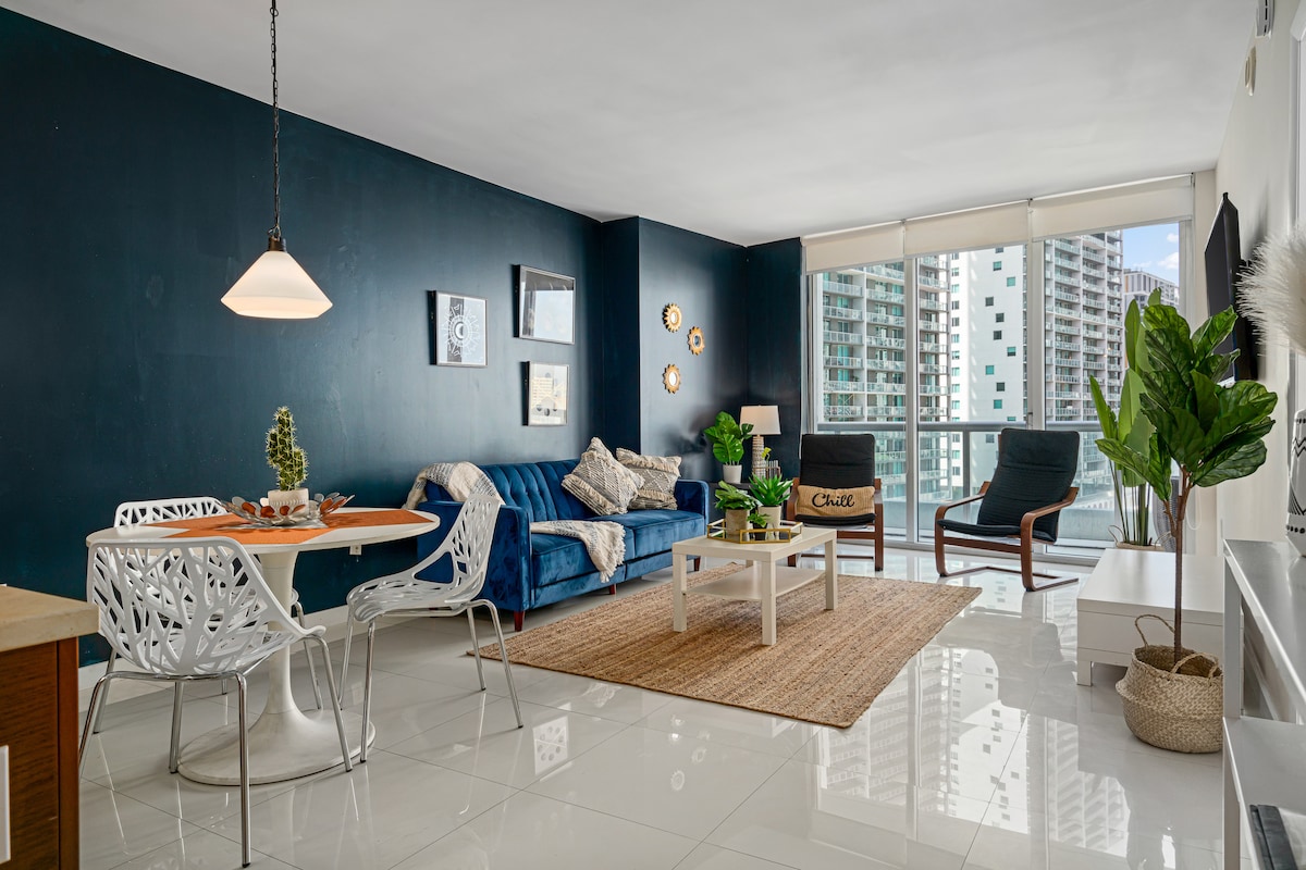 A bright living area is displayed, featuring a blue sofa and a white coffee table on a natural fiber rug. Two modern chairs flank the table, and a plant adds a touch of greenery. Large glass doors allow natural light and offer views of the surrounding buildings.