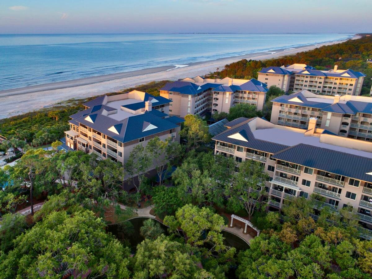 An aerial view showcases the beachfront resort nestled among lush greenery. The coastline extends along a serene beach, while several multi-story buildings are visible, exhibiting modern architecture with blue roofs. The tranquil ocean reflects soft hues of blue and pink from the sky.