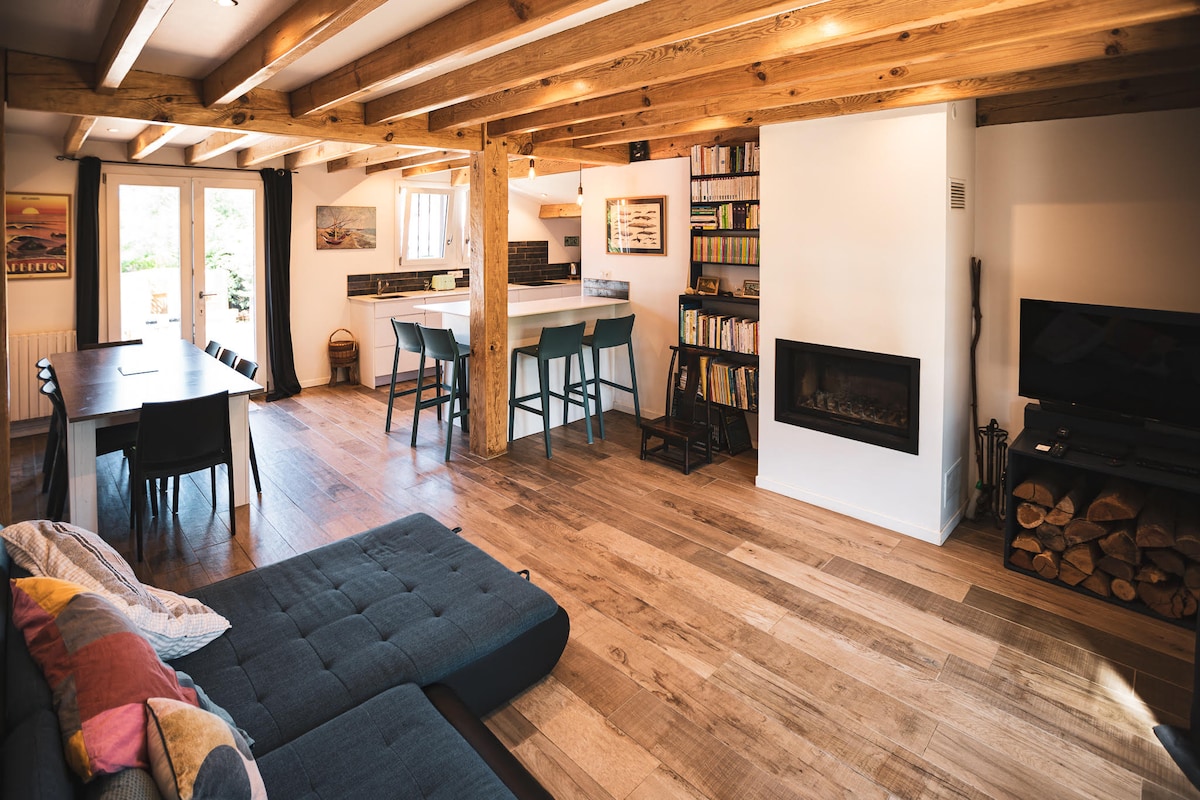 An open living space exhibits wooden beams and a combination of light and dark furnishings. A comfortable sofa is arranged facing a modern fireplace. A dining area features a large table with black chairs, while a kitchen bar with stools offers additional seating. A bookcase occupies one wall.