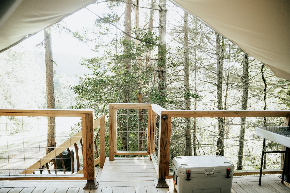 A wooden deck offers views of the surrounding forest, with trees visible in the background. A cooler is positioned on the deck, and a table is placed nearby, providing a space for outdoor dining or preparation.