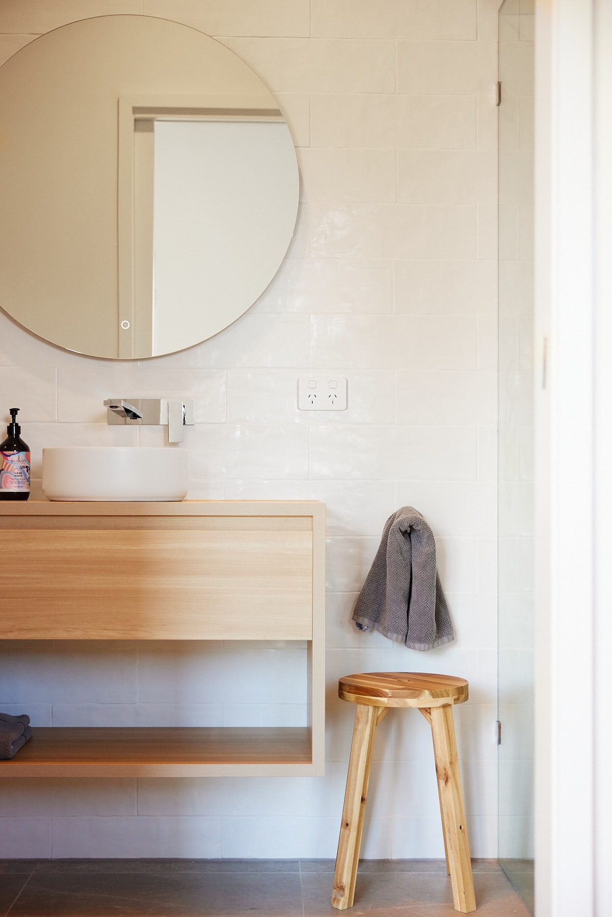 A modern bathroom features a sleek wooden vanity with a round mirror above. A white basin rests on the vanity, while a bamboo stool is positioned nearby. A grey towel hangs from a hook on the wall, and the space is adorned with light-colored tile.