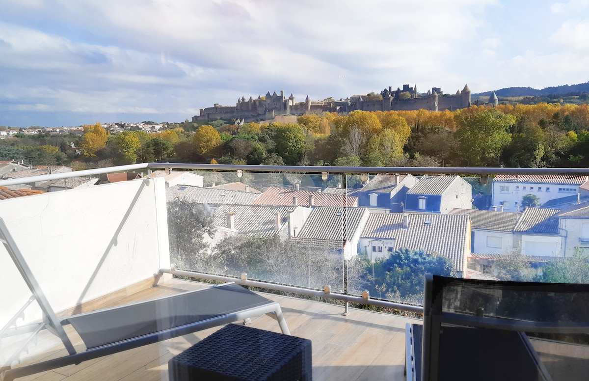 A private terrace features sleek outdoor furniture, including a sun lounger and a small table. The panoramic view overlooks the historic city walls of the Cité Médiévale, framed by greenery and autumn foliage, with houses and rooftops visible below.