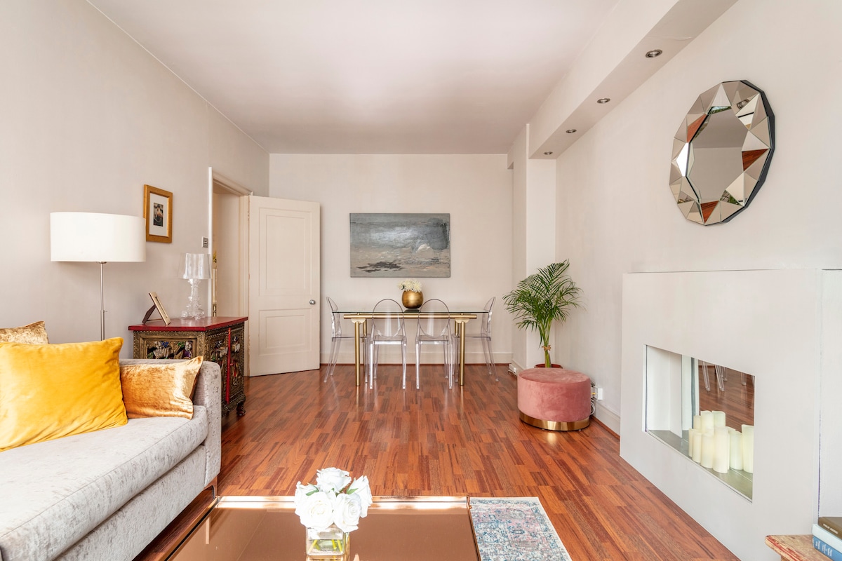 A modern living area features a light grey sofa adorned with yellow cushions, complemented by a decorative coffee table. A dining table with transparent chairs is set against a neutral wall. Natural light fills the space, highlighting a contemporary fireplace and a large mirror.