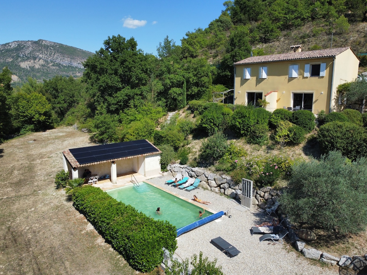 A spacious property is set against a backdrop of rolling hills and greenery. A shared pool is situated in front of the house, surrounded by well-kept landscaping. Lounge chairs are positioned around the pool area, providing relaxation space under the clear blue sky.
