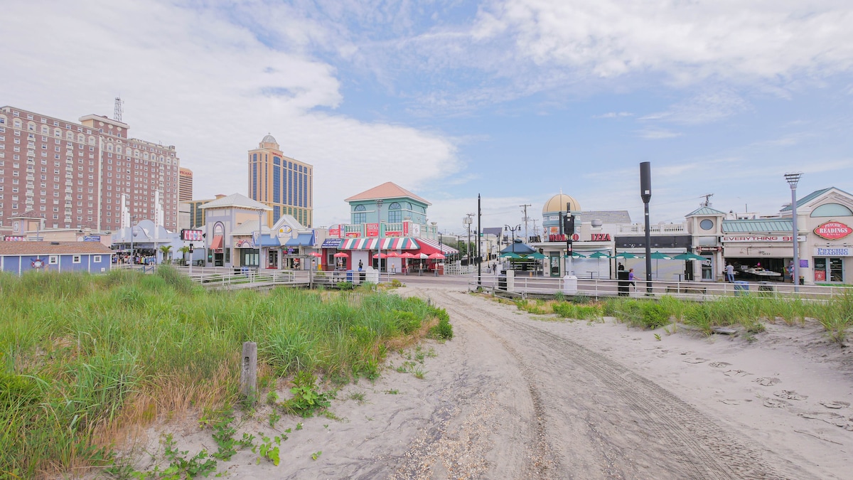 A view of a sandy path leading from grass to a bustling boardwalk, lined with colorful shops and restaurants. Various buildings, including taller hotels in the background, create a lively atmosphere under a partly cloudy sky.