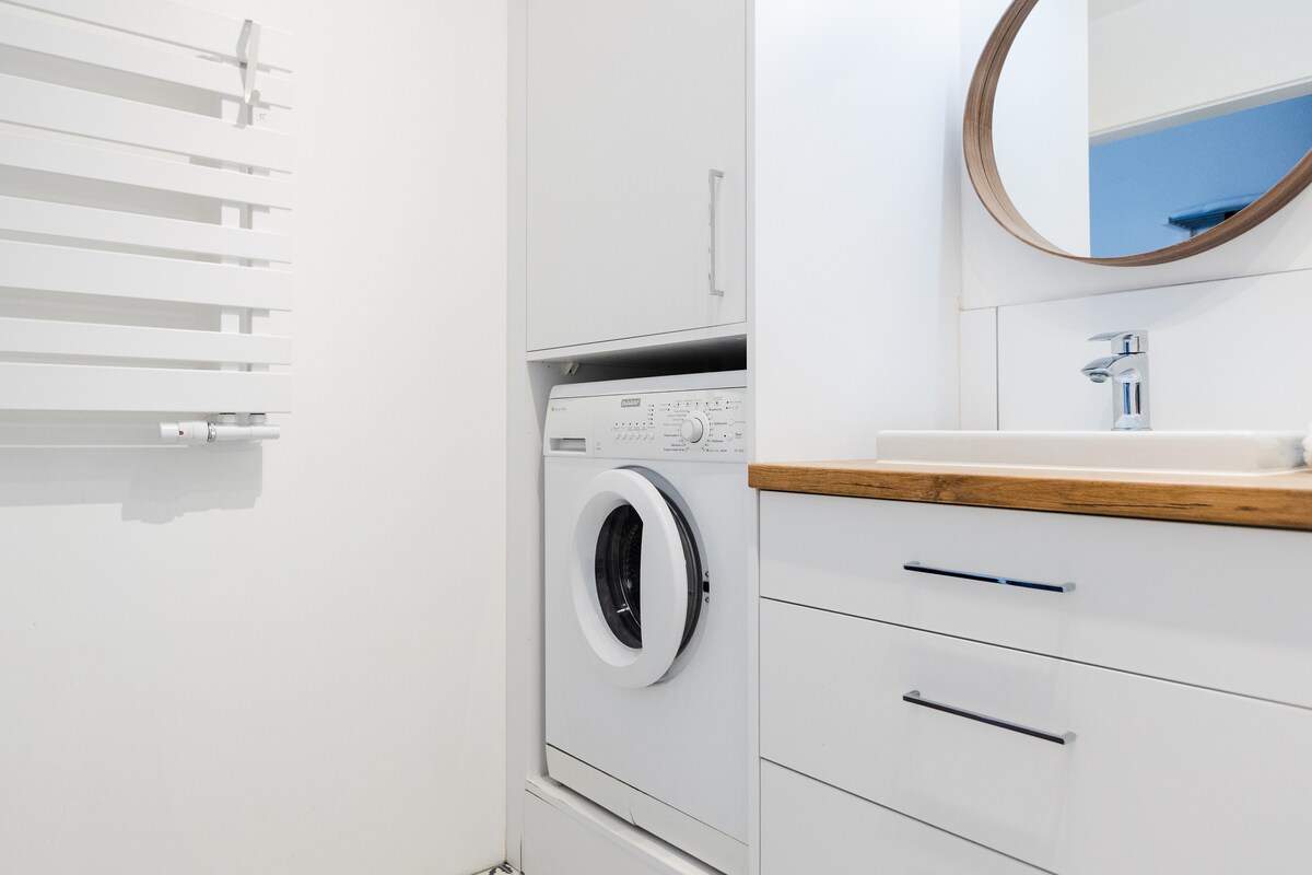 A functional laundry area is visible, featuring a stacked washer and dryer within sleek cabinetry. A round mirror hangs above a minimalist sink, while a wooden countertop adds warmth. A white towel radiator stands on the wall, complementing the modern design.