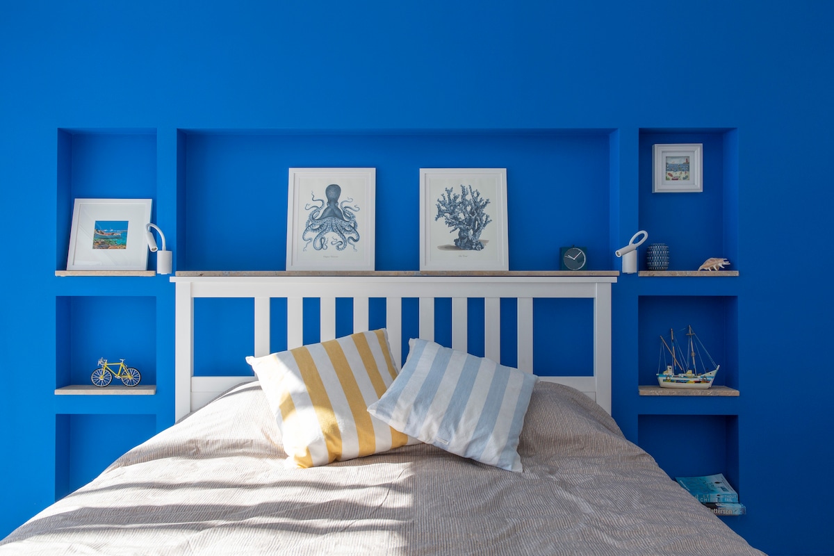 A bright blue wall serves as a backdrop for a white bed featuring two decorative pillows. Shelves are integrated into the headboard, displaying framed artwork and small decorative items. Soft lighting emphasizes the cozy and relaxed ambiance of the room.