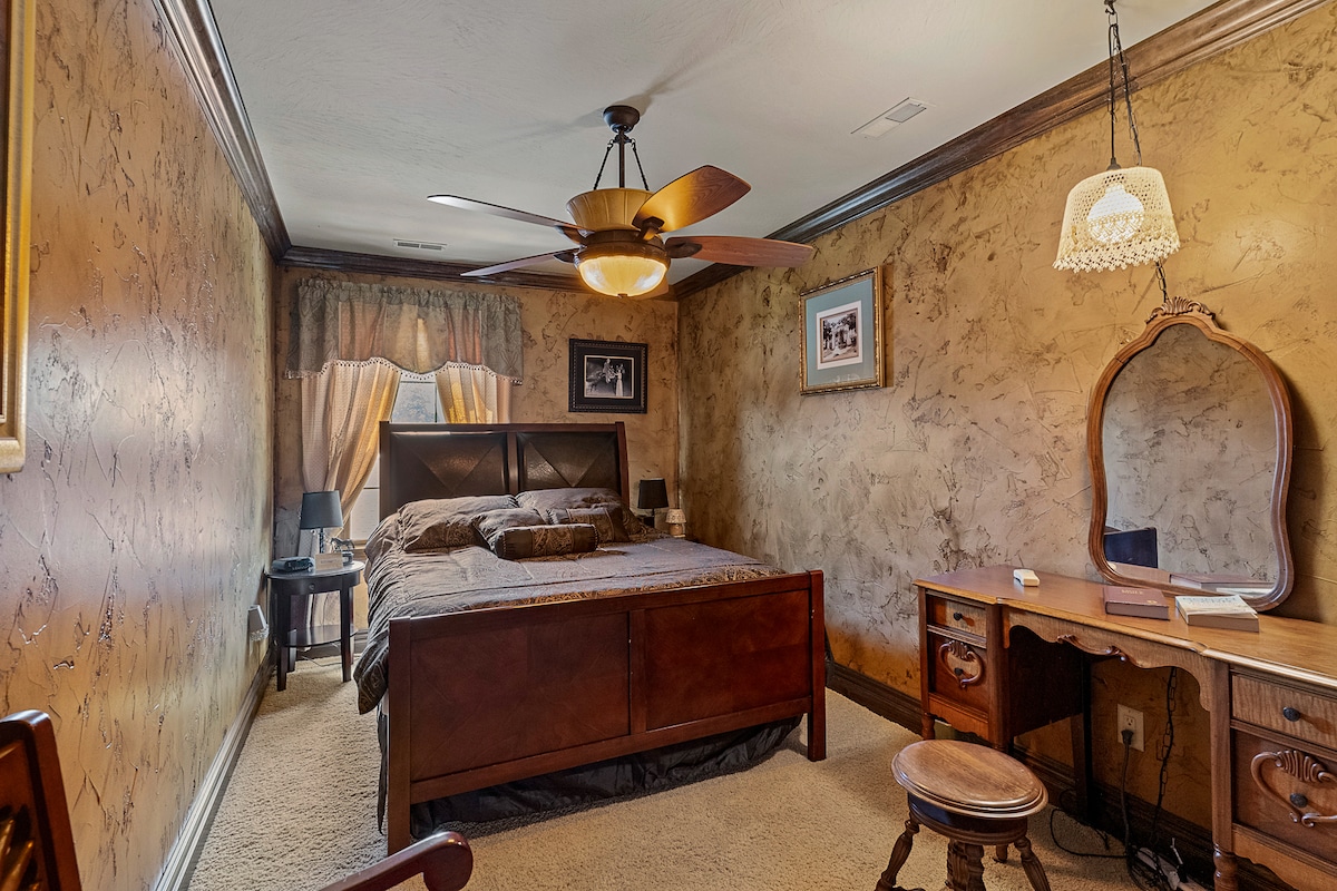 A well-appointed bedroom features a wooden Queen bed with decorative bedding and a ceiling fan overhead. Soft light filters through sheer curtains, illuminating a rustic vanity with a mirror and a stool. Textured walls add warmth to the cozy space.