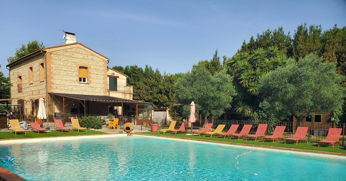 A spacious pool area is surrounded by lounge chairs in various shades of red, creating a serene atmosphere. The two-story building features a traditional stone facade, while lush greenery adds a natural backdrop to the inviting swimming pool.