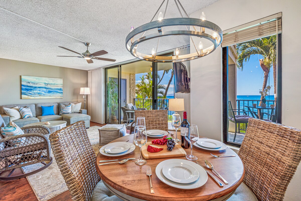 An inviting dining area features a round wooden table set for four, complete with glassware and a platter of fresh fruit. Nearby, a comfortable seating arrangement is visible, adorned with soft pillows. Large sliding glass doors lead to a balcony showcasing ocean views and tropical plants.