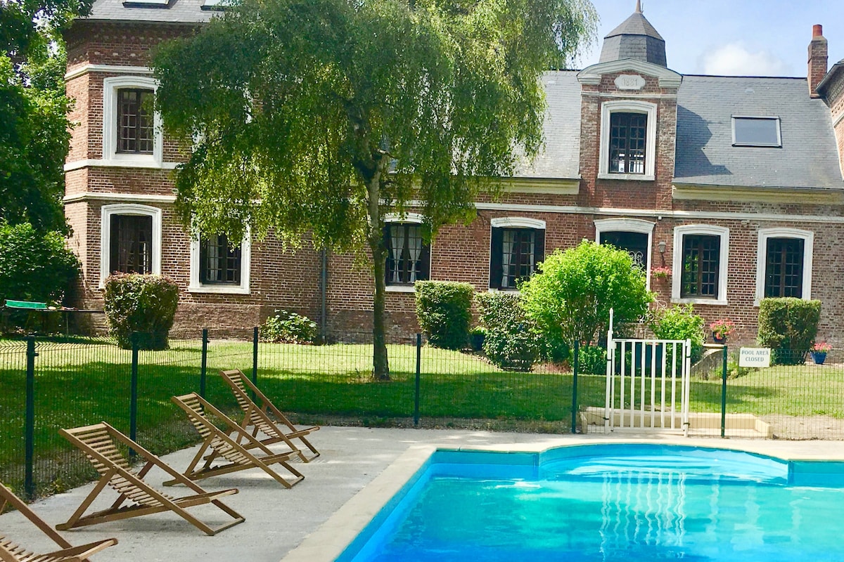 The image shows a heated pool surrounded by a fenced area. Several loungers are arranged neatly along the poolside. In the background, a brick structure features multiple windows and a decorative turret, while well-maintained greenery and shrubs enhance the serene outdoor setting.